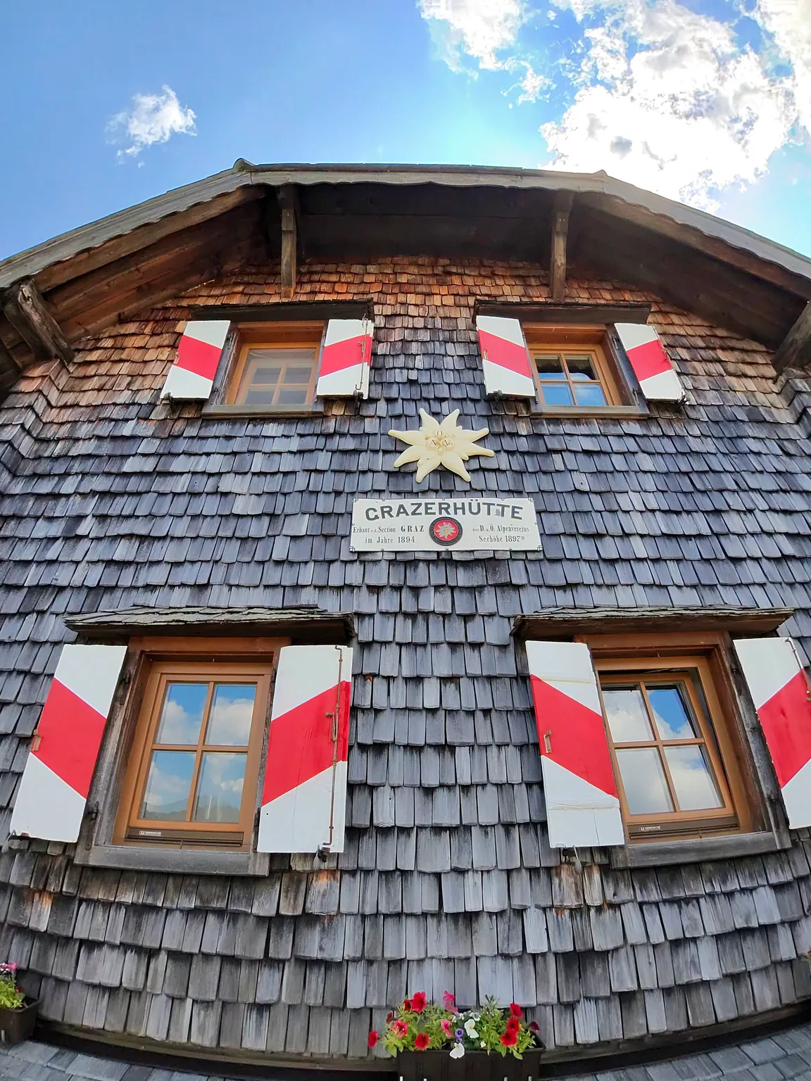Wide-angle view of the wood-shingled Grazer Hütte facade with red-white shutters, an edelweiss emblem and the historic hut sign.