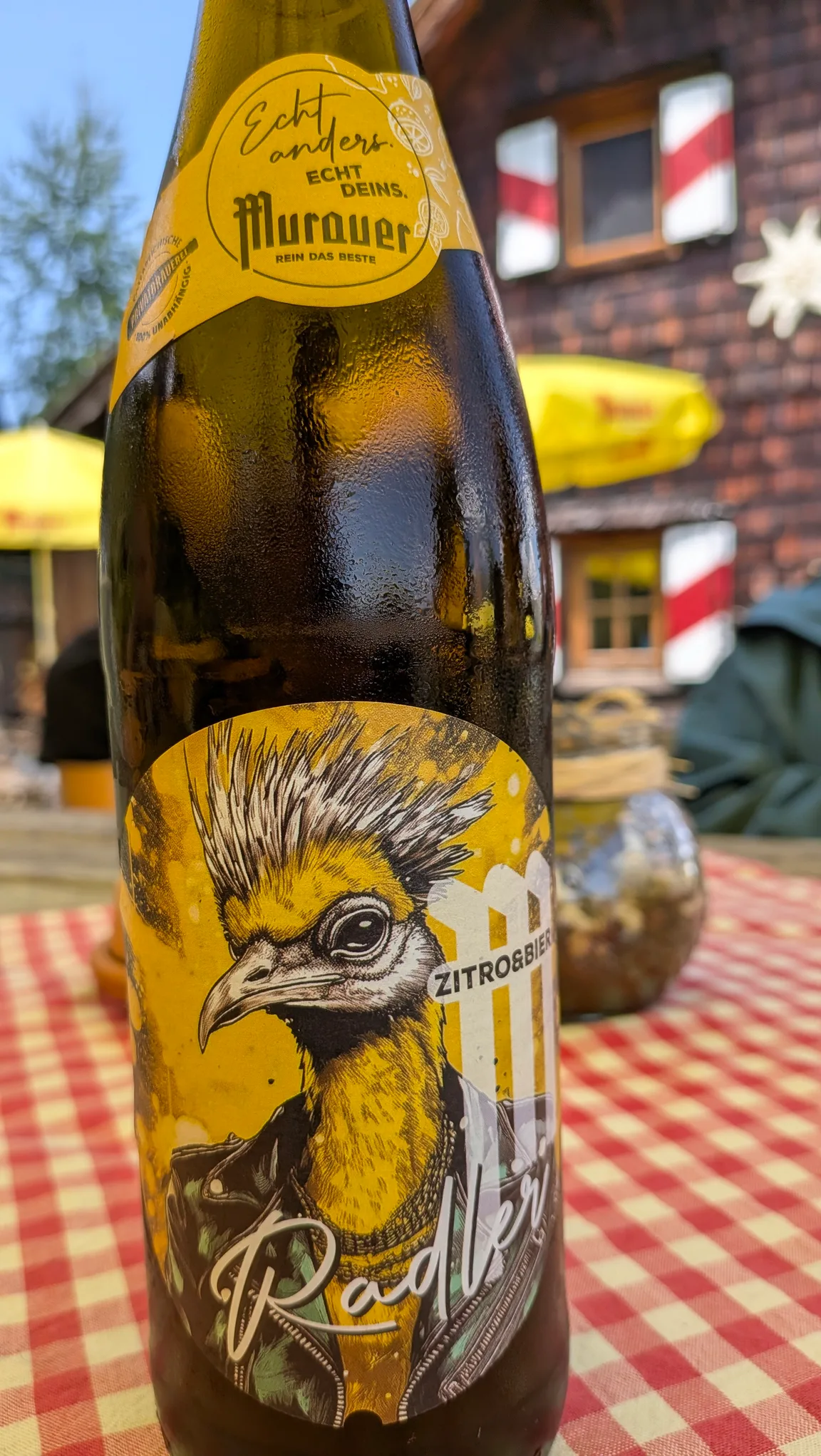 Bottle of Murauer Zitro Radler on a red-checked table on the Grazer Hütte terrace, with the shingled hut facade behind.