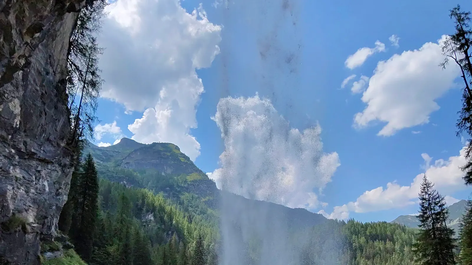View from the secured path beneath the rock overhang, looking out through the falling curtain of the Johanneswasserfall to forested slopes and a rocky peak under a summer sky.