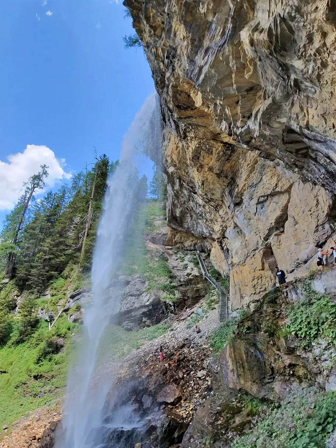 The Johanneswasserfall pouring off a rock overhang, with wooden steps and a railed path climbing the cliff beside it; a few hikers are small figures on the path.